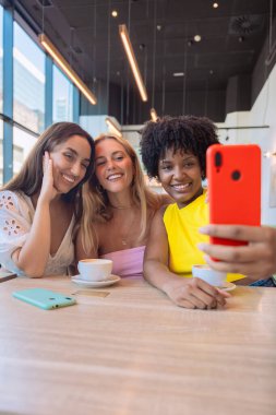 Bir kafede gülümseyen ve poz veren üç farklı kadın. Birlikte kahve içerek eğleniyorlar, akıllı telefonlarıyla eğlenceli bir anı yakalıyorlar. Parlak, neşeli ve sıradan bir ortam.