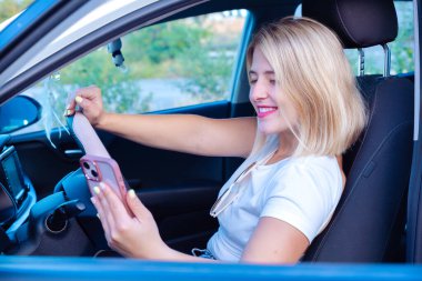 Arabada oturan genç bir kadın cep telefonuyla yan pencereden bakarken neşeyle gülümsüyor..