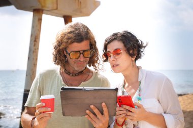 Sahilde oturan orta yaşlı bir adam ve kadın şaşırmış suratlarıyla, akıllı telefonları ve tablet uygulamalarını tarıyorlar. Kavram: Tatiller ve Teknoloji