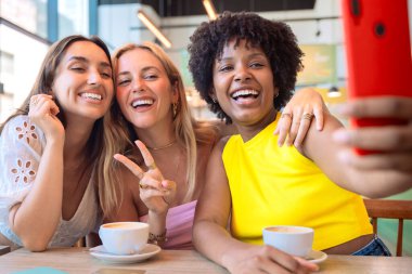 Bir kafede gülümseyen ve poz veren üç farklı kadın. Birlikte kahve içerek eğleniyorlar, akıllı telefonlarıyla eğlenceli bir anı yakalıyorlar. Parlak, neşeli ve sıradan bir ortam.