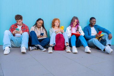 Beş gülümseyen genç öğrenciden oluşan çeşitli bir grup yerde oturup, mavi bir arkaplanda akıllı telefonlarıyla gülümsüyor ve etkileşime giriyorlar..