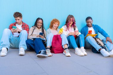 Beş gülümseyen genç öğrenciden oluşan çeşitli bir grup yerde oturup, mavi bir arkaplanda akıllı telefonlarıyla gülümsüyor ve etkileşime giriyorlar..