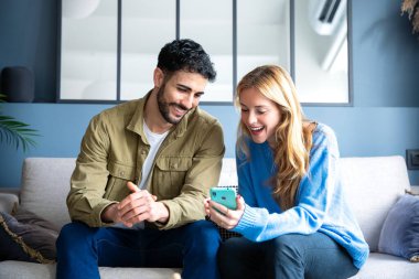  Genç, gülümseyen, çok kültürlü bir çift evdeki koltukta oturuyor cep telefonuyla bir cihazdaki kablosuz internet uygulamasını tarıyor. Teknoloji konsepti.