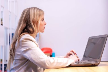 Açık renkli spor ceket giyen profesyonel bir kadın parlak bir ofisteki dizüstü bilgisayarını kullanarak ahşap bir masada oturuyor. Ayarlar renkli nesneler gibi ince arkaplan ayrıntılarını içerir.