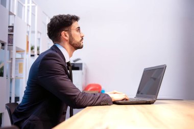 Açık renkli blazer ceket giyen profesyonel bir adam parlak bir ofisteki bir dizüstü bilgisayarla ahşap bir masada oturuyor. Ayarlar renkli nesneler gibi ince arkaplan ayrıntılarını içerir.