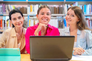 Üç genç kadın bir kütüphanede dizüstü bilgisayar ve tabletle birlikte oturuyorlar. Etrafları kitaplıklarla çevrili ve rahat bir akademik ortamda ortak bir öğrenme ya da çalışma oturumu izlenimi veriyorlar.
