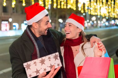 Noel Baba şapkalı mutlu çift, bayram boyunca süslenmiş bir sokakta hediyelerini ve kahkahalarını paylaşıyor ve bayram anlarının tadını çıkarıyor..