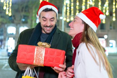 Noel Baba şapkası takan mutlu bir çift dışarıda hediyelerini değiş tokuş ediyor ve bayram ruhunun tadını çıkarıyor. Parlayan ışıklarla çevrili bir kahkahayı paylaşan adam güzel bir hediye tutuyor..