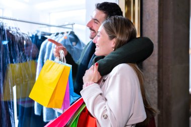 Gülümseyen bir çift mağaza vitrinine bakarken alışveriş yapmaktan hoşlanır. Kadın renkli alışveriş torbalarını tutarken erkek elbiseyi işaret eder. Sahne neşe, beraberlik ve tatil alışverişini gösteriyor.