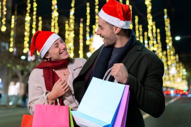  Noel Baba şapkası takan, Noel ışıklarının altında duran gülümseyen bir çift. Birbirlerine sıcak bir şekilde bakıyorlar, neşeli bir tatil ortamı yaratıyorlar. Biri telefonu tutuyor, anıları yakalıyor..