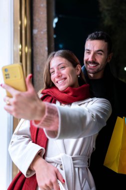 Şenlikli bir şehir gecesinde ellerinde renkli alışveriş torbalarıyla selfie çeken gülümseyen bir çift. Noel, teknoloji, alışveriş ve bayram kutlamaları için mükemmel.