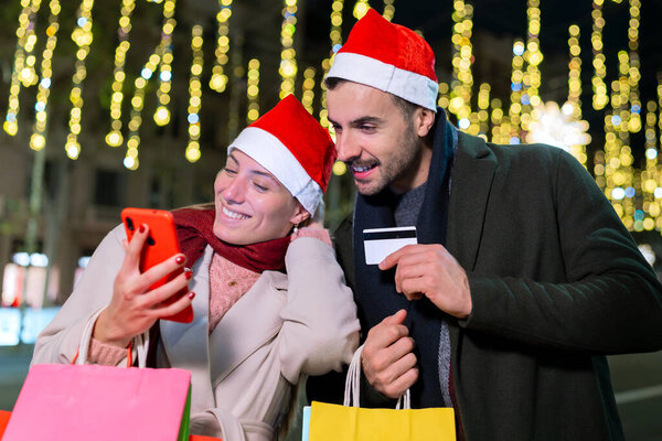 Веселая пара магазинов в Интернете, используя смартфон и кредитную карту при светящихся праздничных огней. Женщина держит сумки с покупками, пока они наслаждаются праздничной атмосферой на открытом воздухе в городской обстановке