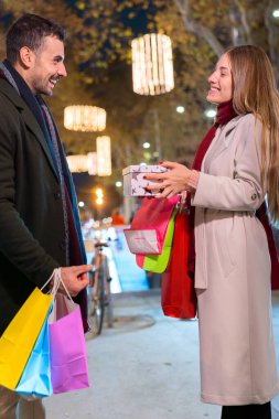 Şenlikli bir şehir gecesinde ellerinde alışveriş torbalarıyla hediye kutusunu değiş tokuş eden heyecanlı çift. Noel, bayram kutlaması, hediye verme ve şehir ortamında neşeli anlar için mükemmel.