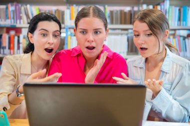 Kütüphanedeki üç kadın dizüstü bilgisayar izlerken şaşırdı. Sahne, kitaplıklar ve çalışma materyalleri ile çevrili şaşkınlık, merak veya şok duygularını taşır.