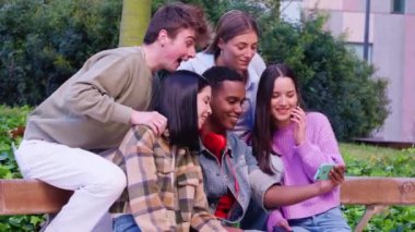 Bir bankta oturan beş farklı genç arkadaştan oluşan bir grup canlı bir açık hava ortamında akıllı bir telefonla gülüp selfie çekiyorlar. Neşe, dostluk ve modern sosyal bağları yakalar..