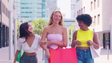 Güneşli bir günün tadını çıkaran üç genç kadın alışveriş torbaları taşıyor ve şehir ortamında yürürken gülümsüyorlar. Arkadaşlık, alışveriş, moda ve yaşam tarzı gibi kavramlar için mükemmel..