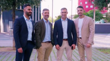 Şehir ortamında gülümseyen ve poz veren dört kendine güvenen iş adamı. Resmi giysiler içinde takım çalışması, profesyonellik ve modern bir iş ortamında başarıyı temsil ediyorlar..