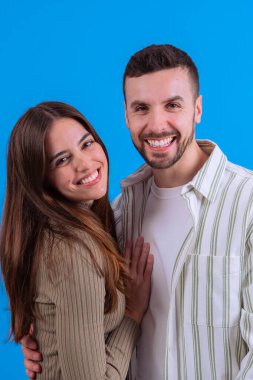 Neşeli genç bir çift kameraya sımsıkı sarılıyor ve gülümsüyor parlak mavi bir arka plana karşı. Aşk, ilişkiler, mutluluk veya pozitif yaşam tarzı kavramları için ideal görüntü.