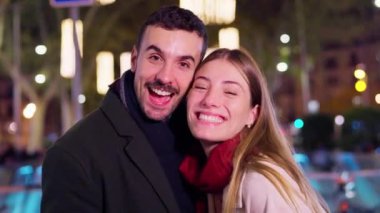 Neşeli bir caddede, parlak Noel ışıklarıyla çevrili gülümseyen bir çift. Kadın renkli alışveriş torbaları tutuyor, Noel alışverişinin ve kutlamaların keyfini çıkarıyor.