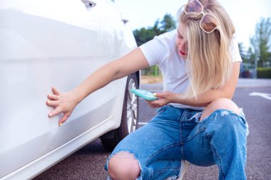 Arabasının yanında diz çöken bir kadın, araç tarafındaki çiziğe bakıyor. Fotoğraf çekmek ya da araba hasarını kontrol etmek için elinde bir akıllı telefon tutuyor. Sahne, detaylara dikkat ve ilgi gösteriyor..