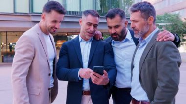 Takım elbiseli dört iş adamından oluşan bir grup modern ofis bölgesinde bir akıllı telefona bakıyorlar, ekranda bir şey tartışıyorlar, takım çalışması, iş ve teknolojiyi temsil ediyorlar.