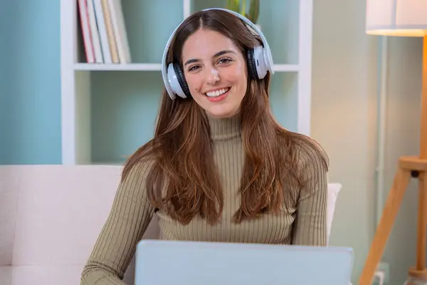 Evde dizüstü bilgisayar ve kulaklıkla çalışan genç bir kadın rahat bir ofis ortamında kameraya gülümsüyor. Uzak çalışma, teknoloji ve üretkenlik kavramları için mükemmel..