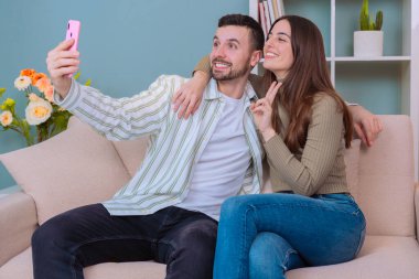 Samimi bir oturma odasında selfie çeken bir çift. Gülümser ve ellerinde akıllı bir telefonla poz verirler. Keyifli ve samimi bir ev ortamında dinlenme anı..