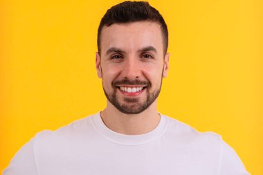 Gülümseyen genç bir adam parlak sarı bir arka planın önünde duruyor, düz beyaz bir tişört giyiyor. Dost canlısı ve kendinden emin bir ifadesi var. Sıradan ve cana yakın bir görünüm sergiliyor..