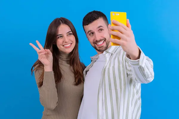 Mavi arka plan önünde akıllı telefonuyla selfie çeken mutlu bir çift. Gülümsüyorlar ve el sallıyorlar. Keyifli bir ortamda birlikte vakit geçiriyorlar..