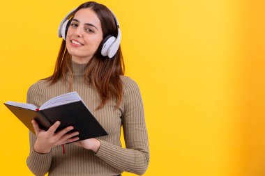 Beyaz kulaklık takan, elinde açık bir kitapla, parlak sarı bir arka plana karşı duran neşeli genç bir kadın. Öğrenme, sesli kitaplar veya eğitici temalar için mükemmel bir resim. Boşluğu kopyala