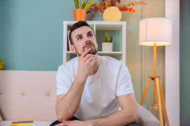 Bir adam derin düşüncelere dalmış, rahat bir oturma odasında oturuyor. Kanepede, çenesinde bir eliyle, bitkilerle ve hafif ışıklandırmayla rahatlamış, yansıması için huzurlu bir atmosfer yaratıyor..