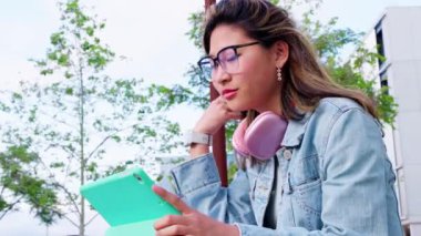 Kot ceketli bir kadın tabletini açık havada kullanmaktan hoşlanıyor, onunla etkileşime girerken gülümsüyor. Yeşillik ve modern mimari ile çevrili pembe kulaklık takıyor..