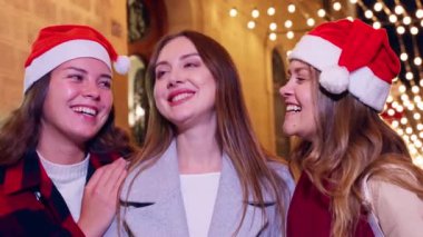 Üç mutlu arkadaş Noel 'i kutluyor, Noel Baba şapkaları takıyor ve festival ışıklarının keyfini çıkarıyorlar. Arka planda parlak tatil dekorasyonlarıyla birlikte dışarıda poz veriyorlar, neşe saçıyorlar..