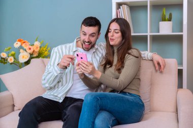 Bir çift rahat bir kanepede oturuyor, birlikte akıllı telefona bakarken keyifli bir anı paylaşıyorlar. Gülümsüyorlar ve ekranı gösteriyorlar, evde eğleniyorlar ve rahatlıyorlar.