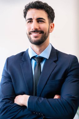 Kolları bağlı ve dostça gülümseyen takım elbiseli kendinden emin bir iş adamı. Parlak bir ofiste duruyor. Profesyonel bir görünümü var. Başarı ve liderlik hissini yansıtıyor.