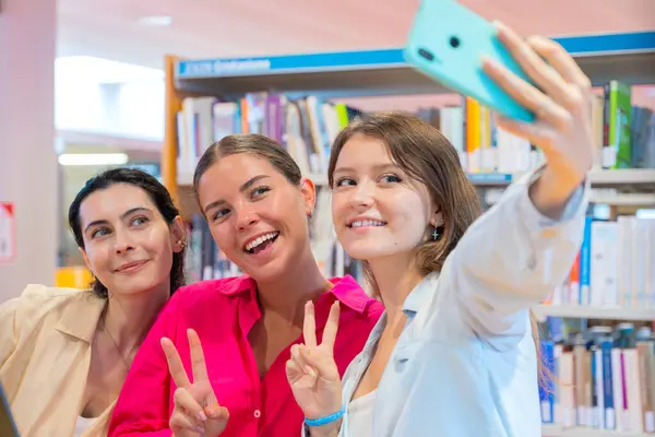 Üç kadın kütüphanede bir selfie için poz veriyor, gülümsüyor ve barış işaretleri gösteriyor. Kitap rafları arka planda, canlı ve eğlenceli bir atmosfer yaratıyorlar..