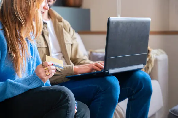Bir çift birlikte kanepede oturuyor, online alışveriş için dizüstü bilgisayar kullanıyorlar. Bir kişi kredi kartını tutarken diğeri laptopla etkileşime geçip rahatlamış ve modern bir alışveriş deneyimi yaratıyor..