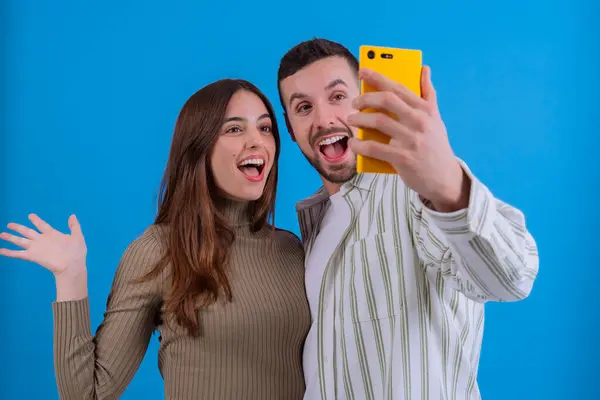 Mavi arka plan önünde akıllı telefonuyla selfie çeken mutlu bir çift. Gülümsüyorlar ve el sallıyorlar. Keyifli bir ortamda birlikte vakit geçiriyorlar..
