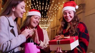 Noel Baba şapkası takan üç kadın, festival ışıklarıyla süslenmiş bir caddede gülüp hediyelerini değiş tokuş ediyorlar. Neşeli an, tatil sezonunun ve arkadaşlığın ruhunu yakalar..