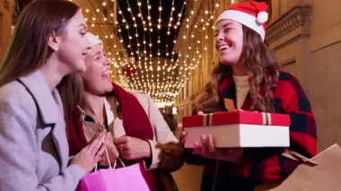 Noel Baba şapkası takan üç kadın, festival ışıklarıyla süslenmiş bir caddede gülüp hediyelerini değiş tokuş ediyorlar. Neşeli an, tatil sezonunun ve arkadaşlığın ruhunu yakalar..