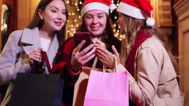 Üç kadın Noel Baba şapkası takıyor, gülümsüyor ve telefonla gülüyor, ellerinde alışveriş torbalarıyla bir festival caddesinde. Sahne, tatil sezonunun neşe ve heyecanını yakalıyor..