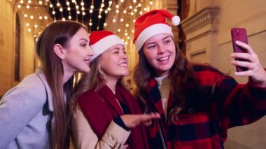 Noel Baba şapkası takan üç neşeli kadın, tatil sezonunda güzel Noel ışıklarının önünde gülümserek selfie çekiyorlar. Birlikte keyifli bir an geçiriyorlar. Şenlikli şehirde kutlama yapıyorlar..