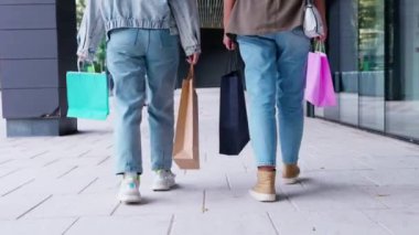 Başarılı bir alışveriş çılgınlığından sonra ellerinde renkli alışveriş torbalarıyla sokakta yan yana yürüyen kadınlar. Sıradan giyiniyorlar, kot pantolon ve spor ayakkabı giyiyorlar, açık hava alışverişinin tadını çıkarıyorlar.