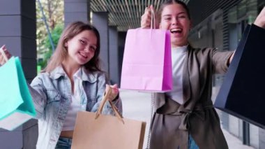 İki mutlu kadın modern bir binanın dışında renkli alışveriş torbaları tutuyor, gülümsüyor ve günlerinin tadını çıkarıyorlar. Sahne, şehir ortamında alışveriş yapmanın heyecanı ve neşesini vurguluyor..