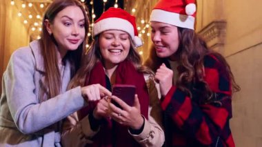 Üç kadın Noel Baba şapkası takıyor, gülümsüyor ve telefonla gülüyor, ellerinde alışveriş torbalarıyla bir festival caddesinde. Sahne, tatil sezonunun neşe ve heyecanını yakalıyor..
