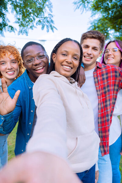 A diverse group of friends takes a joyful selfie outdoors. They are smiling, waving, and enjoying their time together in a bright, sunny environment, showcasing their friendship and positivity.