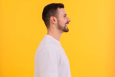 Gülümseyen genç bir adam parlak sarı bir arka planın önünde duruyor, düz beyaz bir tişört giyiyor. Dost canlısı ve kendinden emin bir ifadesi var. Sıradan ve cana yakın bir görünüm sergiliyor..
