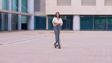  Kadın modern bir şehir bölgesinde scooter kullanıyor. Sıradan giyinmiş ve gezintiden zevk alırken gülümsüyor. Arka planda canlı şehir atmosferini gösteren çağdaş binalar yer alıyor..