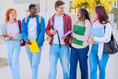 Bir grup öğrenci dışarıda birlikte yürürken gülümsüyor. Ellerinde defterler ve klasörler tutuyorlar, gündelik kıyafetler giyiyorlar ve parlak, modern bir ortamda vakit geçiriyorlar..