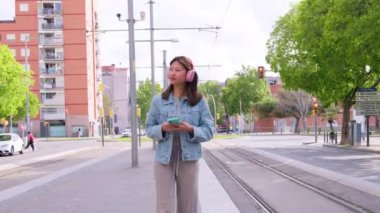 Pembe kulaklık ve kot ceket takan şık bir genç kadın elinde akıllı bir telefonla tramvay raylarında güvenle yürüyor. Şehir arkaplanı canlı ve modern bir yaşam tarzını yansıtıyor.